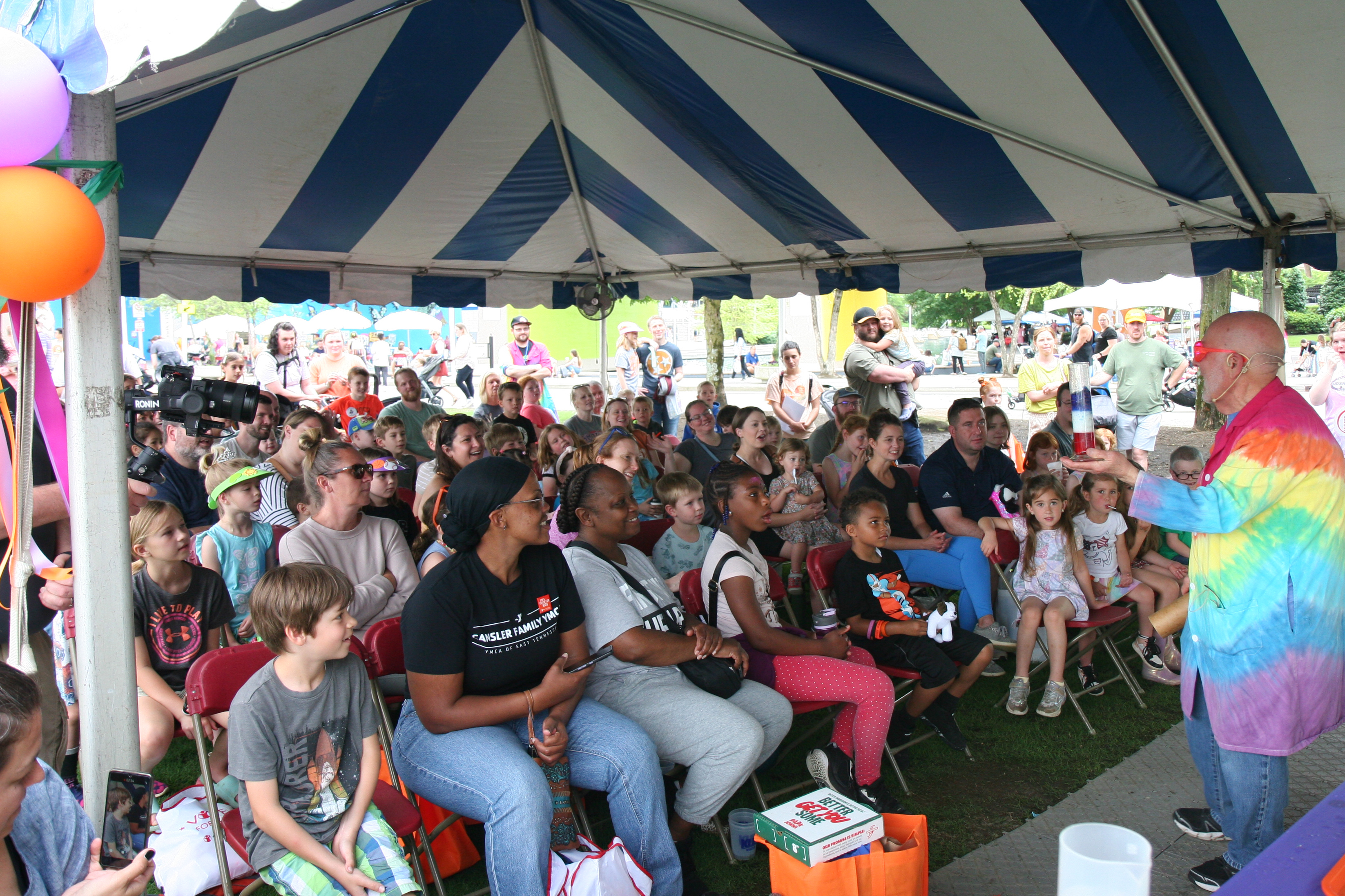 presenter in tie-dye coat enthralls a crowd seated under a tent