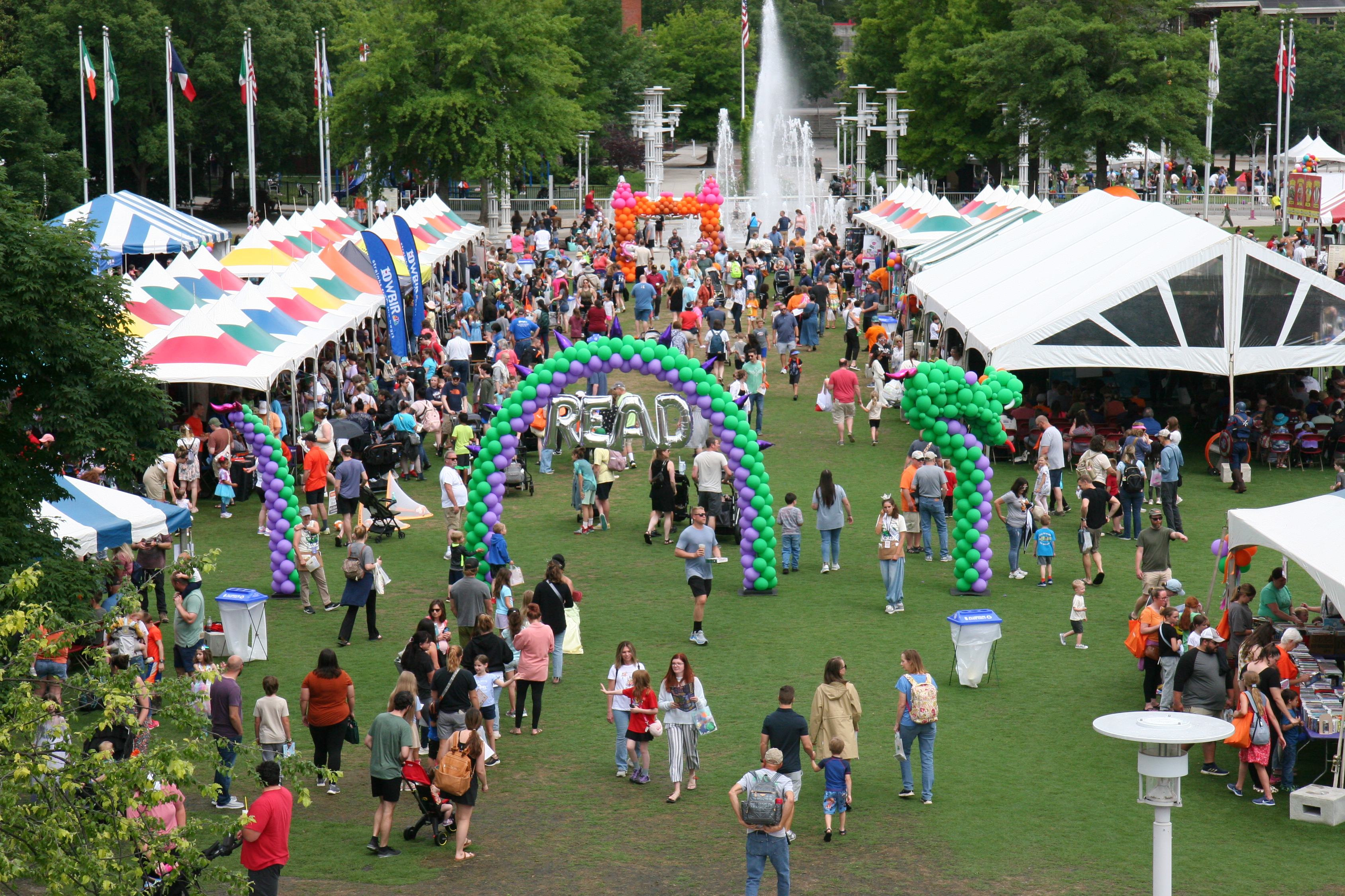 overview photo of outdoor festival with colorful tents, dragon balloon arch, large crowd, and fountain in the background