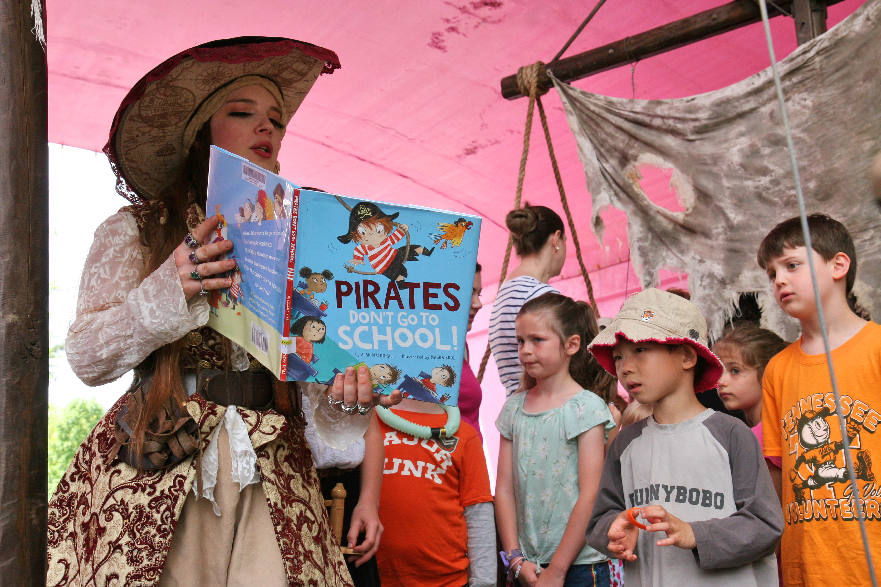 woman dressed as pirate reads a book to children with a mast and sail in background