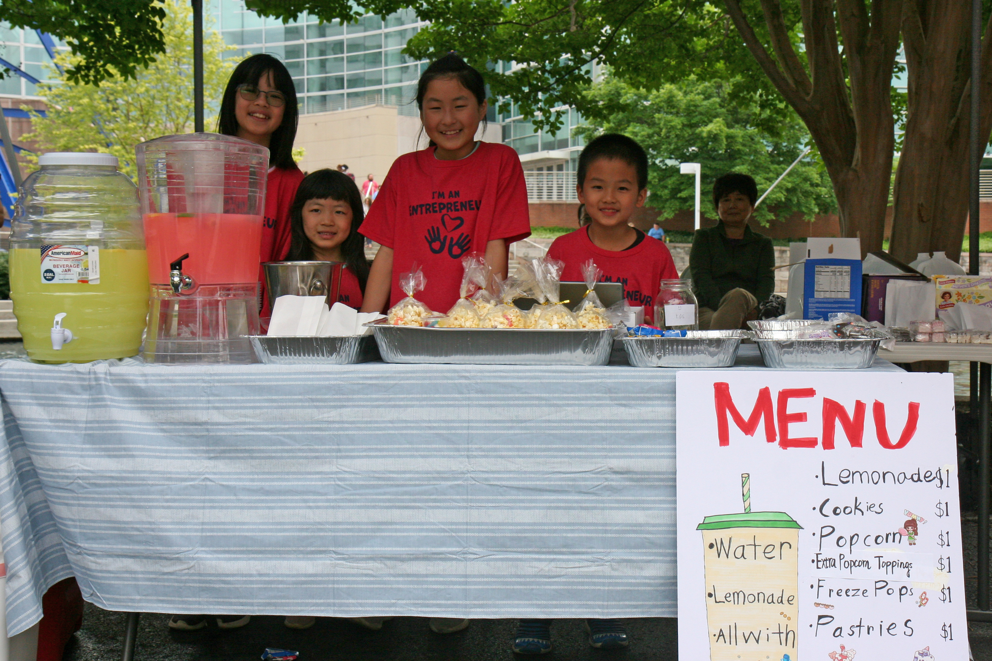 four kids stand behind a table selling lemonade with hand-drawn Menu poster in front