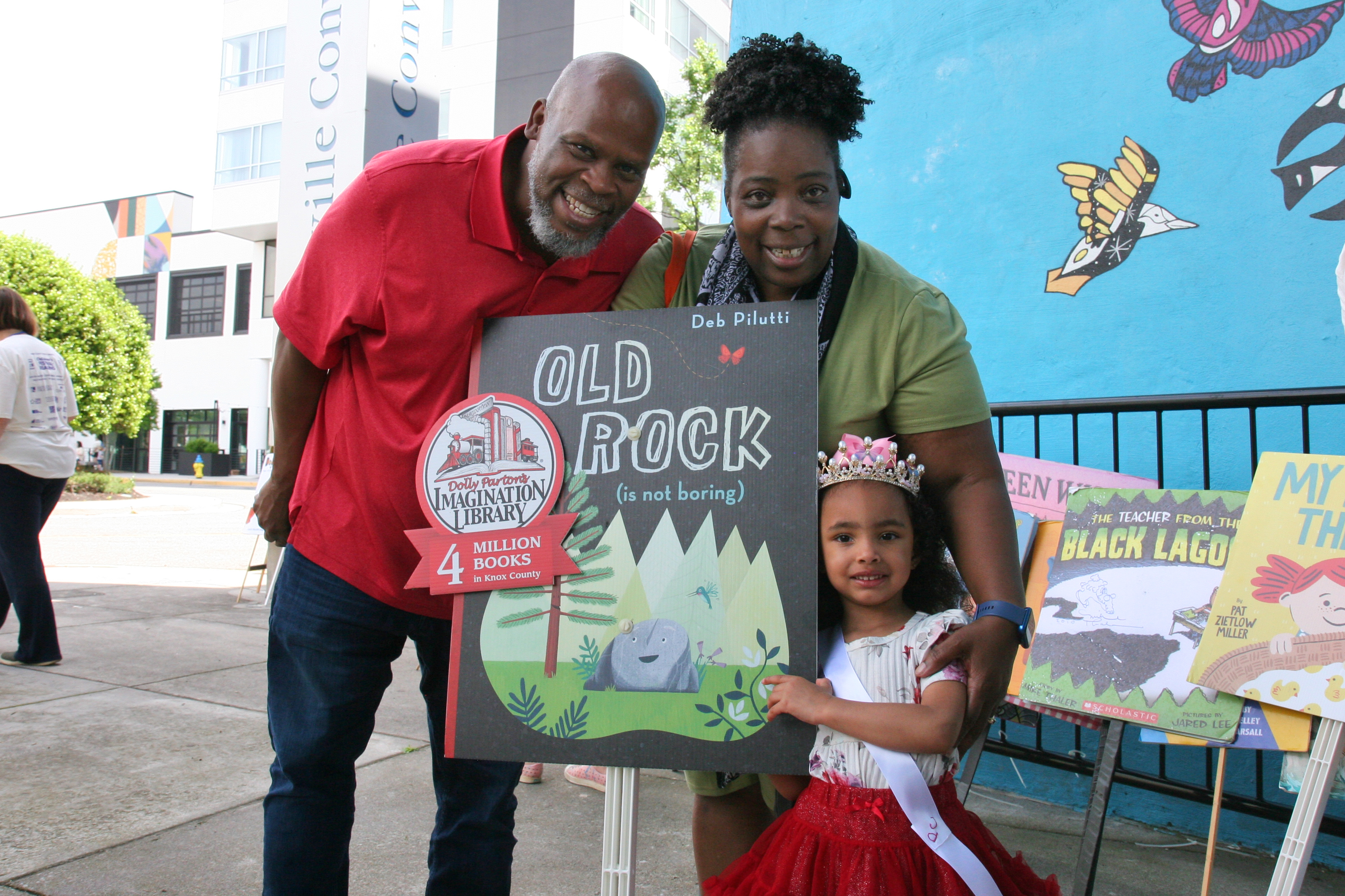 young Black girl wears a tiara and holds a sign of the book Old Rock with her smiling grandparents