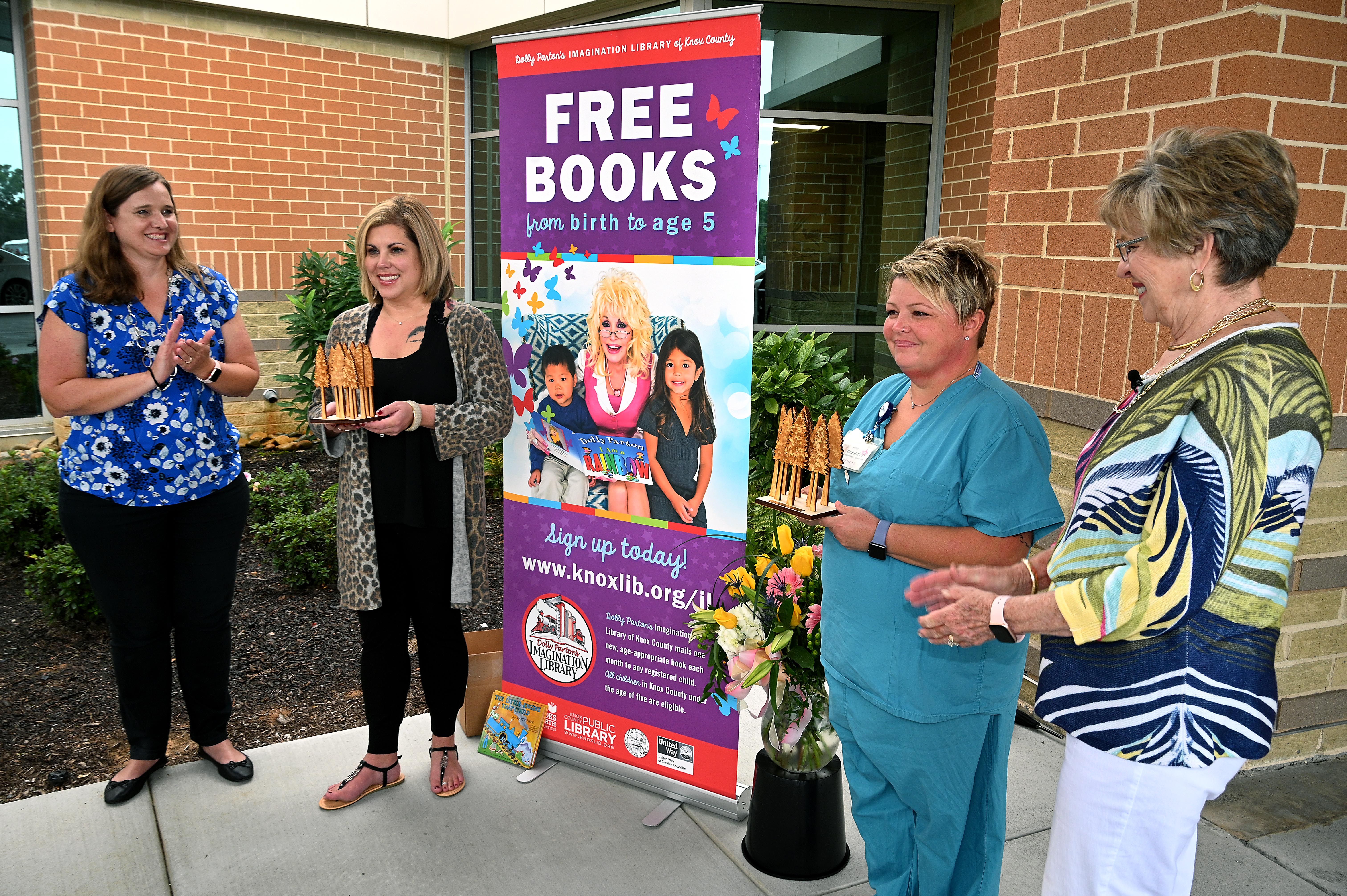 Danielle Velez, Holly Woodlee, Christy Richardson, and Bonny Naugher stand in front of a brightly-colored Imagination Library sign at the presentation