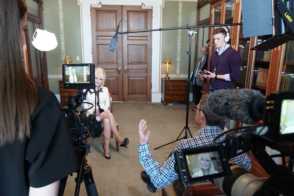 Dolly Parton, seated, is being interviewed for the documentary by a camera crew