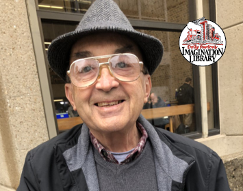 Pastor John, a slim older gentleman with glasses and a gray fedora, smiles for the camera.