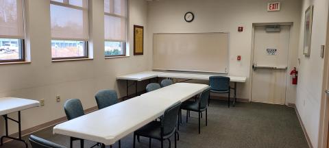 Photo shows a carpeted room with windows along one wall; white plastic tables and cushioned chairs are set up.