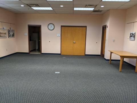 Photo shows rear of a carpeted room with entry door, closet doors, and kitchen door. Along right wall is a wooden table. No chairs are set out. Projection screen and dry-erase board on opposite wall are not shown.