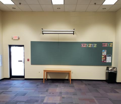 Photo shows carpeted room with outlet in center of floor, outer door, projection screen, wooden table, garbage can.